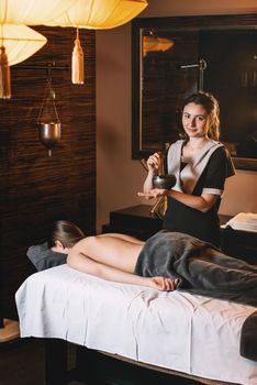 Specialist holding in hands Brass Hammer Singing Bowl. Young charming girl on a panchakarma procedure laying on a massage table. beautiful woman spending time at modern spa cabinet relaxing. Soft yellow light.