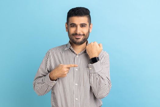 Cheerful positive businessman with beard pointing at wristwatch on hand and smiling, showing new smart clock, wearing striped shirt. Indoor studio shot isolated on blue background.