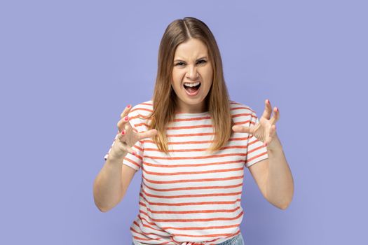 Portrait of angry young adult blond woman wearing striped T-shirt expressing aggressive emotions, raised arms, screaming with hate and anger. Indoor studio shot isolated on purple background.