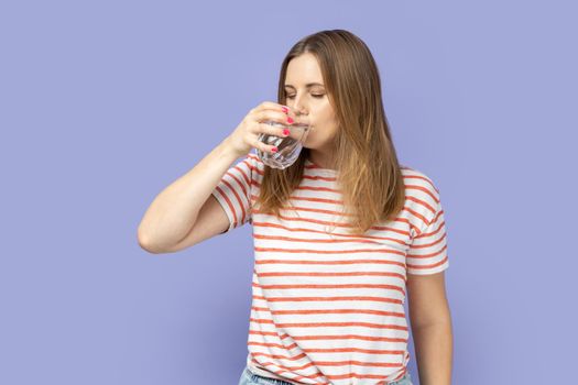Portrait of thirsty attractive beautiful young adult blond woman wearing striped T-shirt drinking water from glass, healthy eating. Indoor studio shot isolated on purple background.