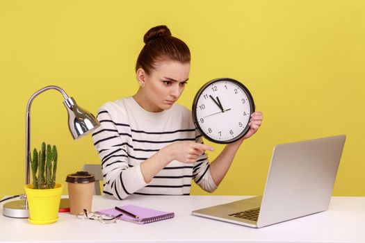Bossy strict woman office worker holding big wall clock, time management, pointing to laptop screen with serious expression. Indoor studio studio shot isolated on yellow background.