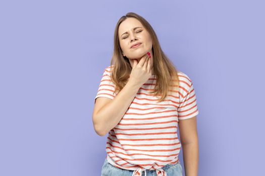 Portrait of sick blond woman wearing striped T-shirt touching her neck, suffering sore throat, viral infection or flu symptoms. Indoor studio shot isolated on purple background.