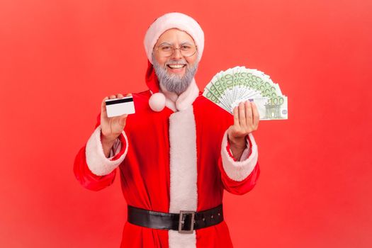 Happy smiling satisfied elderly man with gray beard wearing santa claus costume holding showing fan of euro banknotes and credit card in hands. Indoor studio shot isolated on red background.