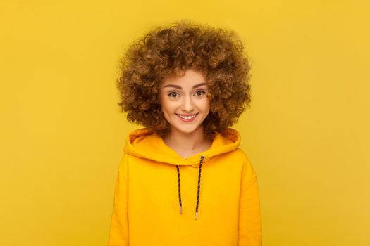 Portrait of pretty smiling happy woman with Afro hairstyle looking at camera with cute positive face, being in good mood, wearing casual style hoodie. Indoor studio shot isolated on yellow background.