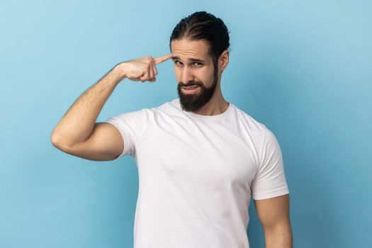 This is insane idea. Portrait of man with beard showing stupid gesture with finger at her temple, displeased with reckless silly joke, out of mind. Indoor studio shot isolated on blue background.