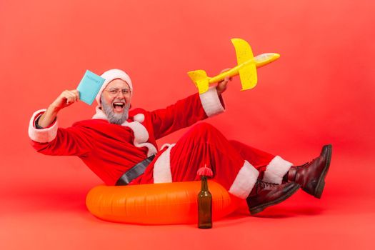 Excited elderly man with gray beard wearing santa claus costume sitting on rubber ring and holding paper plane and passport, enjoying winter traveling. Indoor studio shot isolated on red background.