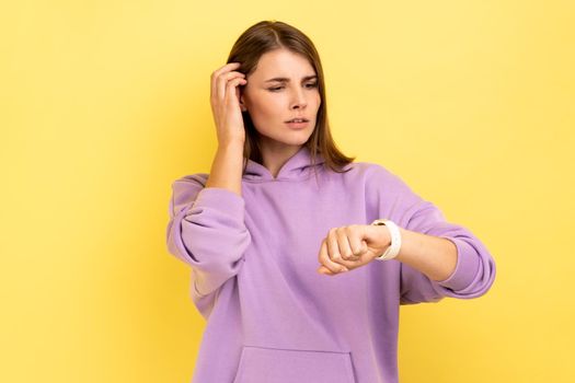 Portrait of pretty woman looking at wrist watch making appointment, meeting time, touching her hair with hand, wearing purple hoodie. Indoor studio shot isolated on yellow background.