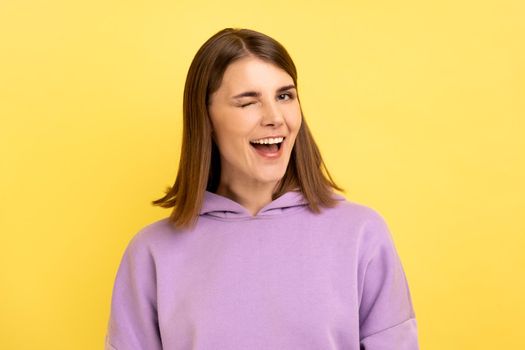 Portrait of positive optimistic woman standing and winking playfully, positive expression, looking at camera, wearing purple hoodie. Indoor studio shot isolated on yellow background.