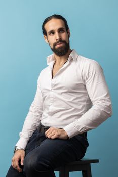 Unsmiling serious dark haired attractive man with beard wearing white shirt and black trousers, looking away with attentive look while sitting on chair. Indoor studio shot isolated on blue background.