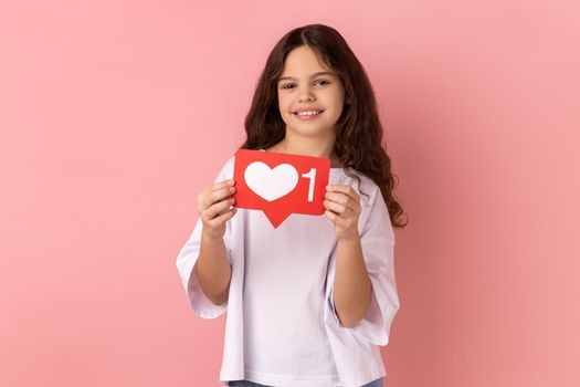 Portrait of little girl wearing white T-shirt holding social media heart Like icon and smiling to camera, recommending to follow and love content. Indoor studio shot isolated on pink background.
