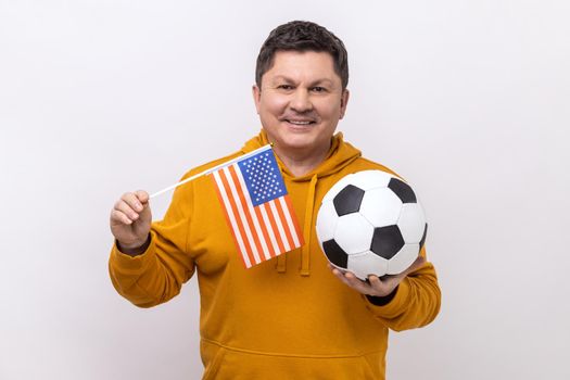 Delighted man holding flag of united states of america and soccer black and white ball, united soccer league, wearing urban style hoodie. Indoor studio shot isolated on white background.