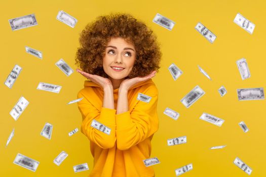 Portrait of cute charming pretty woman with Afro hairstyle wearing casual style hoodie standing in money rain, looking away with angelic face. Indoor studio shot isolated on yellow background.