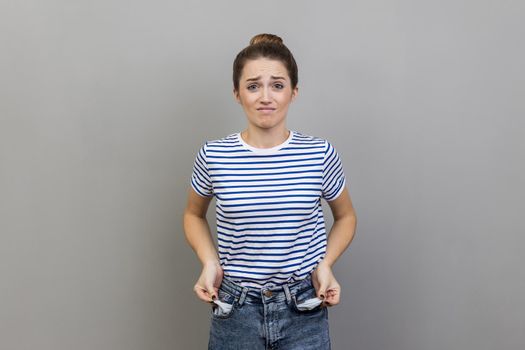 Portrait of upset poor attractive woman wearing striped T-shirt turning out empty pockets, showing no money gesture, worried about debts. Indoor studio shot isolated on gray background.