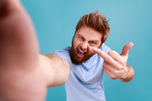 Portrait of bearded man blogger POV, making point of view of photo, showing middle finger to camera, expressing anger and aggressive emotions. Indoor studio shot isolated on blue background.