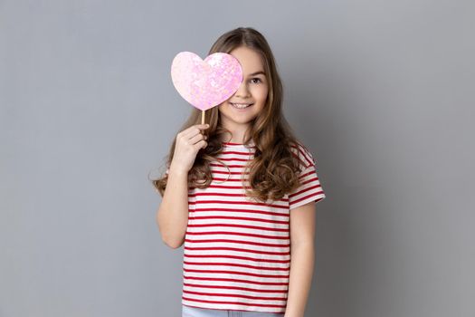 Portrait of satisfied smiling little girl wearing striped T-shirt covering her eye with pink heart on stick, showing romantic feelings. Indoor studio shot isolated on gray background.