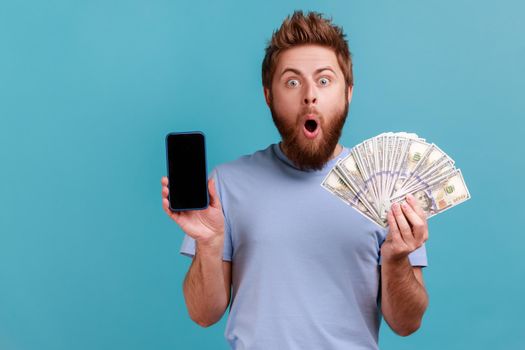 Portrait of surprised amazed handsome bearded man holding dollar banknotes and cell phone with empty screen, online betting, winning. Indoor studio shot isolated on blue background.
