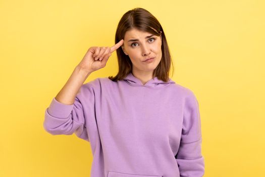 Woman with dark hair showing crazy or stupid gesture holding finger near temple, depressed, has no ideas, wearing purple hoodie. Indoor studio shot isolated on yellow background.