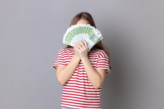 Portrait of unknown anonymous rich little girl wearing striped T-shirt hiding her face behind euro banknotes, income of money. Indoor studio shot isolated on gray background.