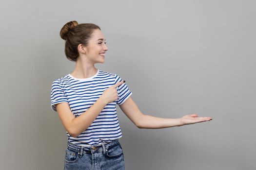 Portrait of satisfied positive happy woman in striped T-shirt presenting copy space on palm, showing empty place for commercial text or goods. Indoor studio shot isolated on gray background.