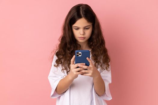 Portrait of serious little girl wearing white T-shirt texting message in social media on cell phone, using mobile network services, chatting online. Indoor studio shot isolated on pink background.