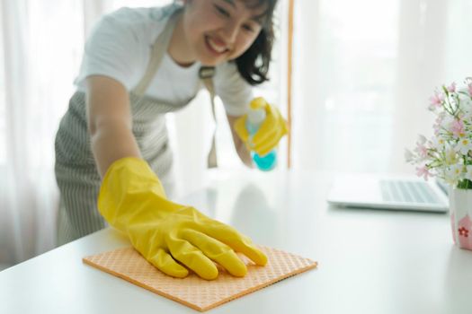 Young woman happily cleaning, wiping, disinfecting table surface using cleaning spray and sponge and wearing yellow protective gloves at home. Woman doing housechores with enjoyment.