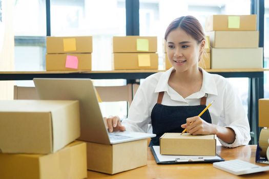 Asian female small online business owner using laptop, packing boxes on table, and checking retail order and client address preparing for shipment. Online business, e-commerce concept.