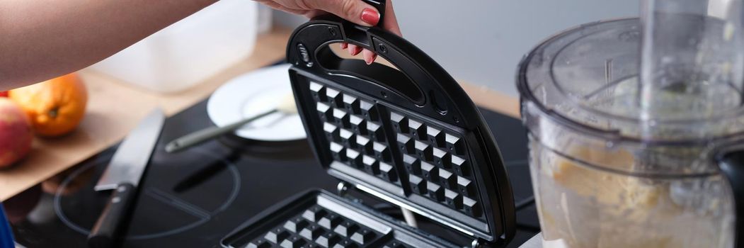 Woman hand opening lid of multi-baker to make viennese waffles near food processor closeup. Household appliances for cooking home baking concept