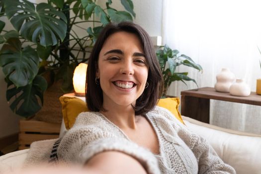 Happy young woman taking selfie at home cozy living room looking at camera. Influencer recording herself with cellphone. Lifestyle and technology.