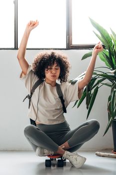 Multiracial young woman sitting on skateboard dancing and listening to music with headphones. Vertical image. Lifestyle concept.