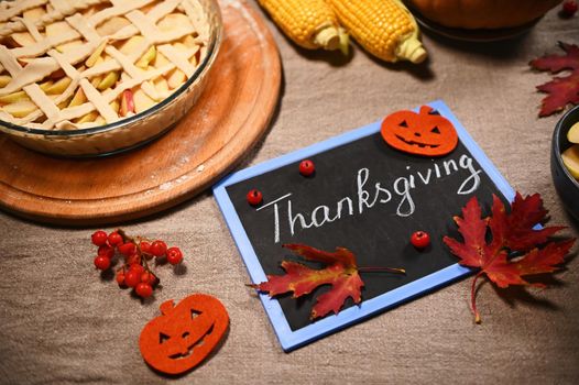 Blackboard with chalk lettering Thanksgiving Day, next to homemade festive pie and autumnal harvest of corn and pumpkin, on a table with linen tablecloth, dry fallen maple leaves and viburnum berries