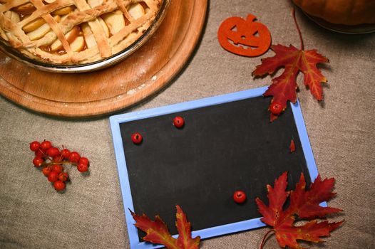 Top view of chalkboard with copy space for advertising text, next to dry autumn maple leaves, viburnum berries, and homemade holiday pumpkin pie with crispy crust for Thanksgiving dinner. Still life