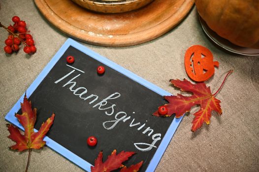 Selective focus on dry red fallen autumnal maple leaves and viburnum berries, lying down next to pumpkins cut from felt, on a chalkboard with lettering Thanksgiving Day. Autumn holidays, Still life