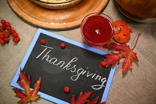 Top view. A cup of hot herbal tea, against blurred background of a pumpkin pie. A blackboard with chalk lettering Thanksgiving Day and dry fallen autumn maple leaves on a linen tablecloth. Still life