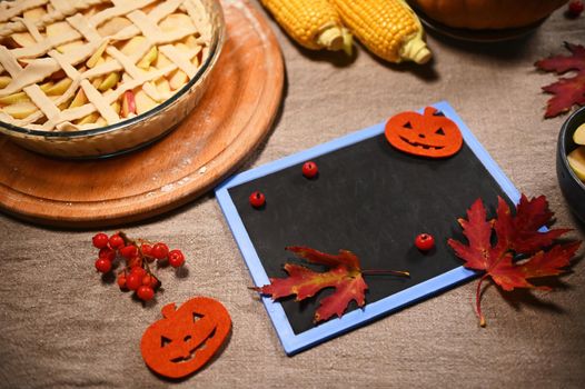 Dry maple leaves, viburnum berries on blank blackboard, copy space for text, harvested corn and freshly baked homemade pumpkin pie with crispy crust lattice, for Thanksgiving Day on linen tablecloth