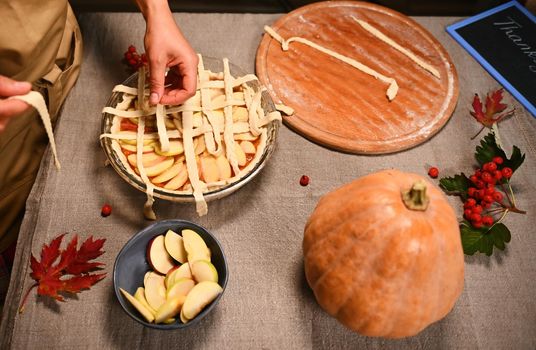 Overhead view of pastry chef in beige apron standing at table with linen tablecloth, decorating homemade pie for Thanksgiving dinner, holding pastry strips, making crusty lattice on top of pumpkin pie