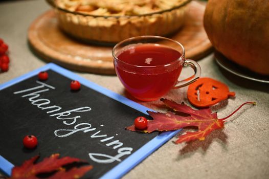 Food still life with a cup of hot herbal tea, against a blurred background of a pumpkin pie. A blackboard with chalk lettering Thanksgiving Day and dry fallen autumn maple leaves on a linen tablecloth