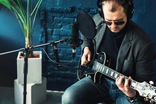 Close up of a man singer in a headphones with a guitar recording a track in a home studio. Man wearing sunglasses, jeans, black shirt and a jacket. side view