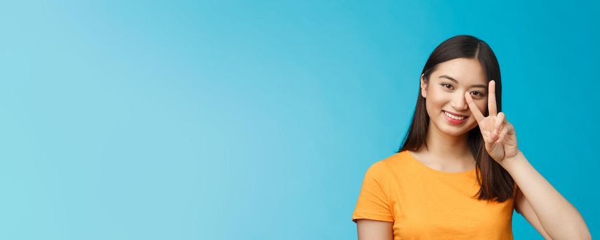 Positive cute and silly asian girlfriend posing photograph lovely smiling, show peace victory sign near face grinning friendly, carefree enthusiastic mood, stand blue background in yellow t-shirt.