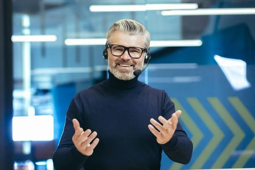 Online business training. A young handsome gray-haired man in headphones with a microphone conducts an online meeting, webinar on camera. He waves his hands, smiles.