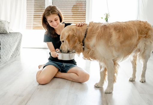 Pretty girl with golden retriever dog sitting on floor at home and holding bowl with water. Beautiful female teenager person with drinking doggy pet in light room