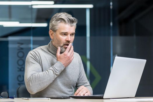 A shocked man, a gray-haired businessman, sits at the desk in the office, works with a laptop. He holds his hand to his mouth, surprised by what he saw on the computer screen.