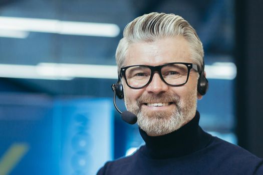 Portrait of a gray-haired handsome man in glasses and a headset with a microphone. Speaker, business trainer, online conference. Looking at the camera, smiling.