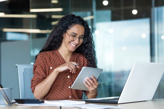 Young beautiful Latin American woman working inside modern office building, paperwork woman uses tablet computer and laptop, accountant calculates reports and accounts, financial documents.