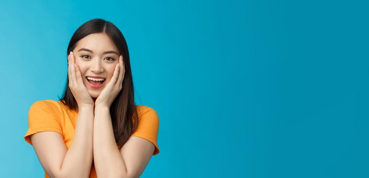 Flirty cute asian girl look surprised, express admiration and joy, touch cheeks, blushing coquettish smiling receive good news, stand blue background happily react amazed and excited.