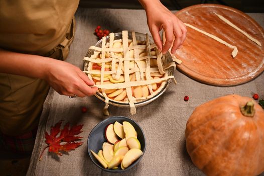 Top view housewife in beige chef's apron standing at table with linen tablecloth, decorating homemade pie for Thanksgiving dinner, holding pastry strips, making crusty lattice on top of pumpkin pie