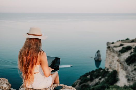 Successful business woman in yellow hat working on laptop by the sea. Pretty lady typing on computer at summer day outdoors. Freelance, travel and holidays concept.
