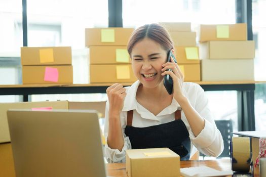 Female small online business owner talking on phone with clients with excitement and delight to recieve and check order online, address preparing to pack order boxes for delivery. Online business concept.