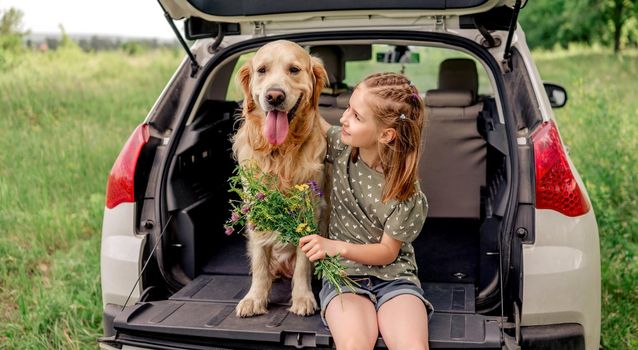 Beautiful little girl sitting with golden retriever dog in the car trunk holding flower bouquet and hugging pet. Child kid with purebred doggy and summer plants in the vehicle at the nature
