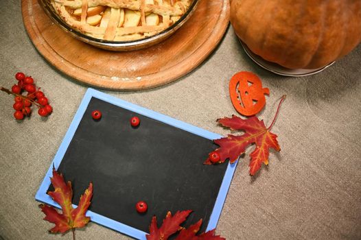 Top view of a blank chalkboard with copy advertising space for promotional text, autumn dry maple leaves, a whole ripe pumpkin and traditional homemade American classic Thanksgiving pie on table