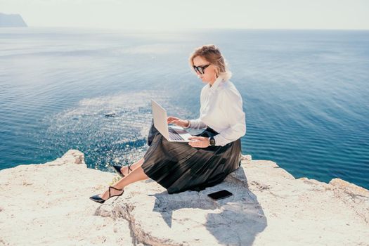 Successful business woman in yellow hat working on laptop by the sea. Pretty lady typing on computer at summer day outdoors. Freelance, travel and holidays concept.
